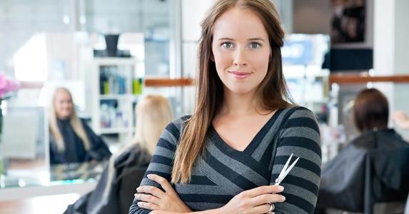 Female hairdresser standing with arms folded with a salon in the background © Tyler Olson/Shutterstock.com