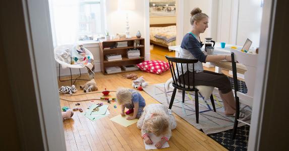 Mother at her home computer with toddlers behind her | Hero Images/Getty Images