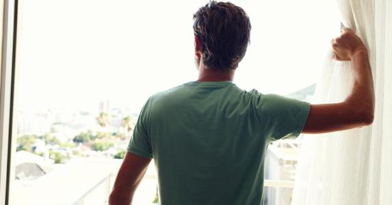 On the fence about buying or building home? Man opening up curtains to look out window | GlobalStock/E+/Getty Images